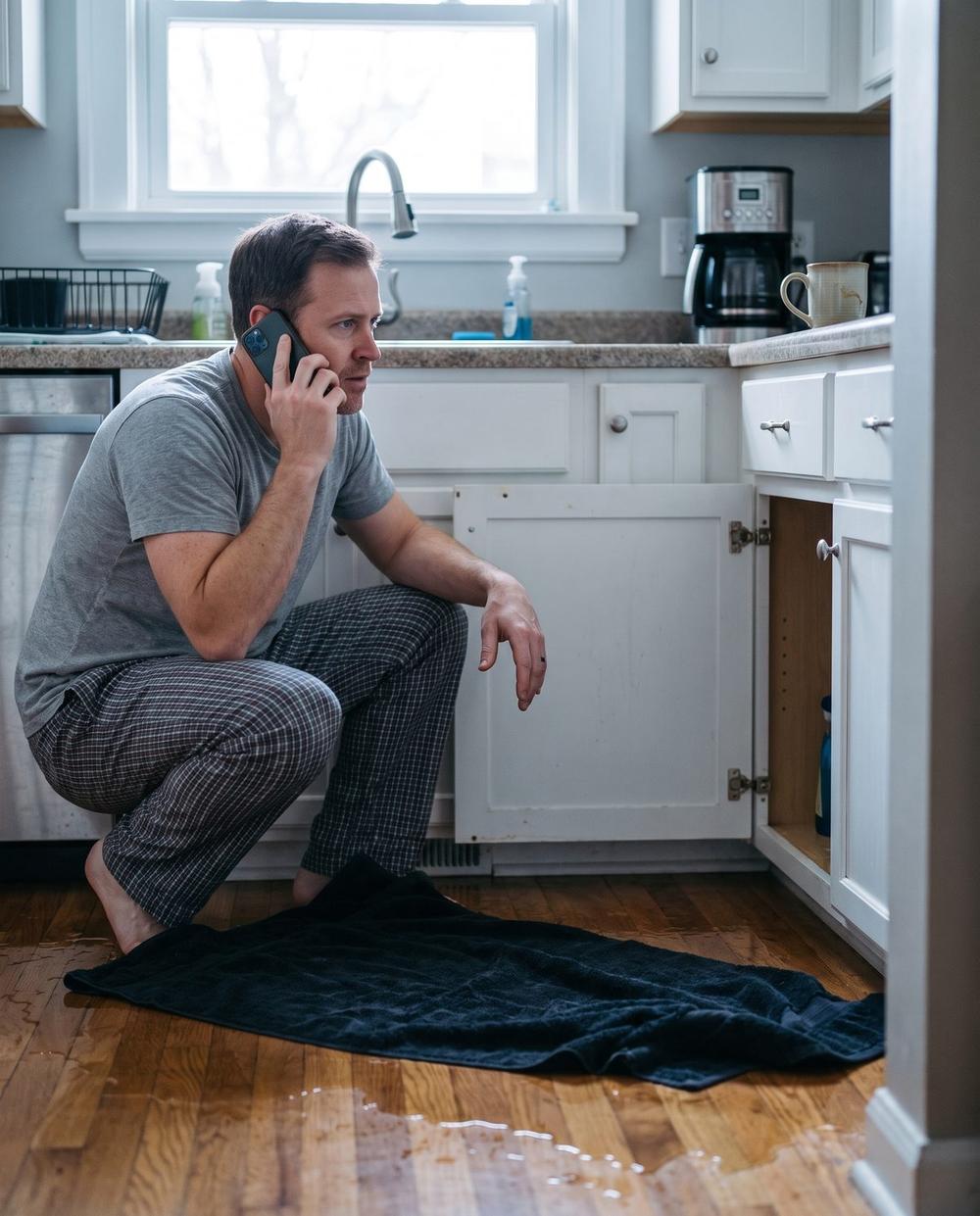 Illustrative image — the same man the next morning, crouched by a leaking kitchen sink with a phone to his ear