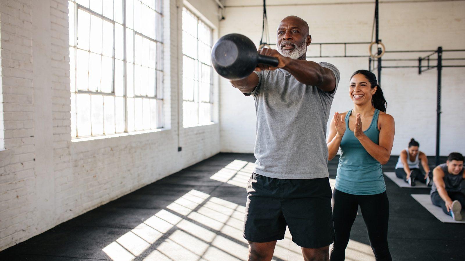 Illustrative image — a man swinging a kettlebell while a trainer cheers him on in a sunlit gym