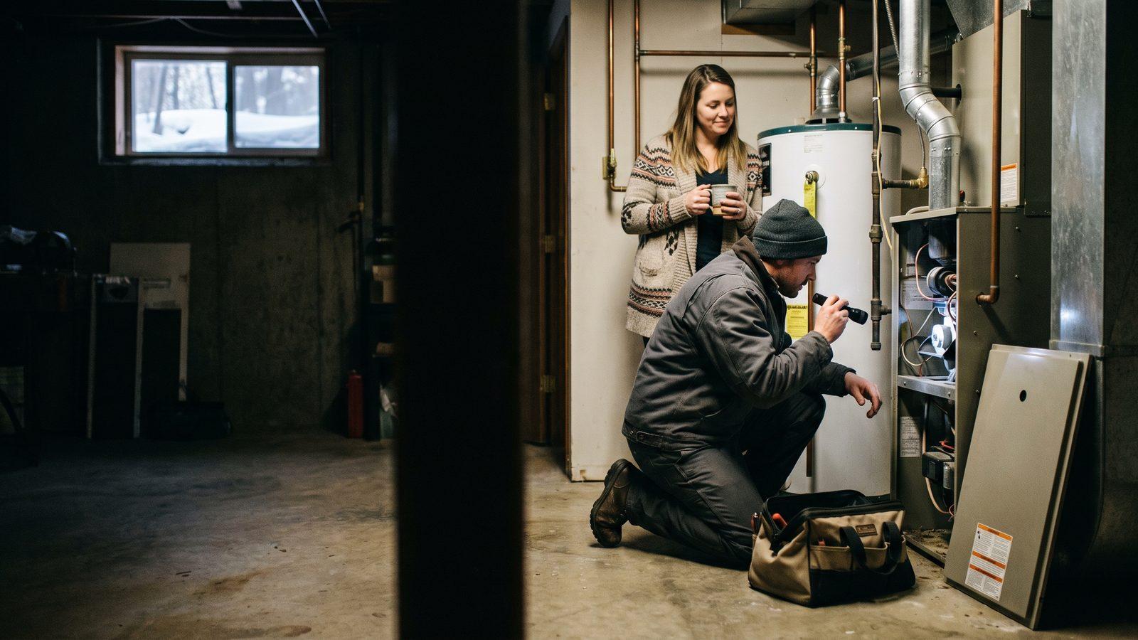 Illustrative image — a technician kneeling at an open furnace with a flashlight in a basement while the homeowner watches with a mug of coffee